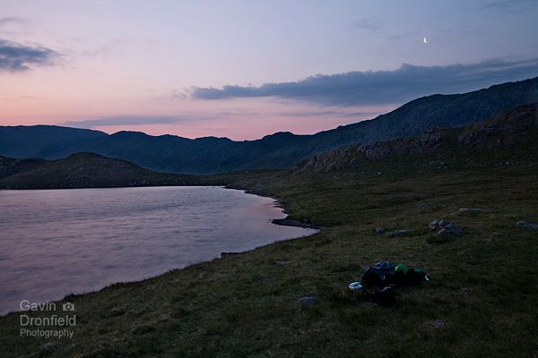 crescent moon in salmon coloured dawn sky over sprinkling tarn with camping gear