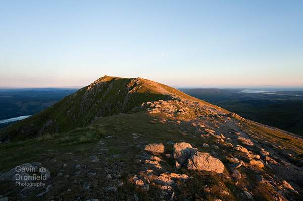 looking along the summit ridge of coniston old man towards summit cairn at sunset with coniston water below