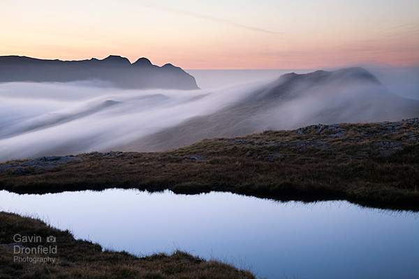 silhouetted langdale pikes above cloud enveloped rossett pike from calm tarn on tongue head under red dawn skies