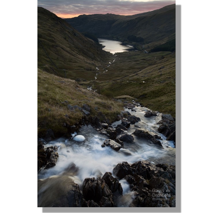 small water beck tumbles over a waterfall flowing towards distant haweswater reservoir under red dawn skies